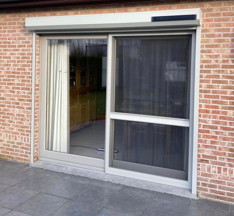 Lichtgrijs zonne-rolluik voor een schuifpui, rolluikkast en geleiders op de gevel.