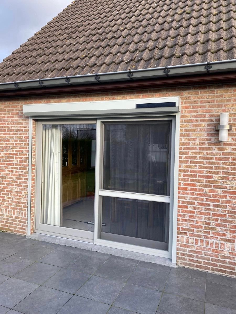 Zonne-rolluik opgebouwd boven een schuifpui, op een huis in baksteen. Het zwarte zonnepaneel zit op de rolluikkast.